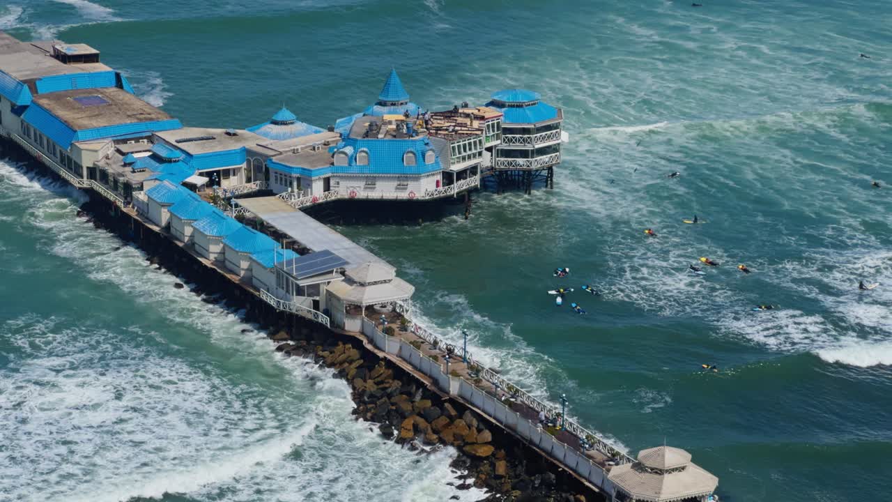 Pier extending into turquoise ocean along La Rosa Nautica Lima Peru from above in tropical weather with surfers catching waves