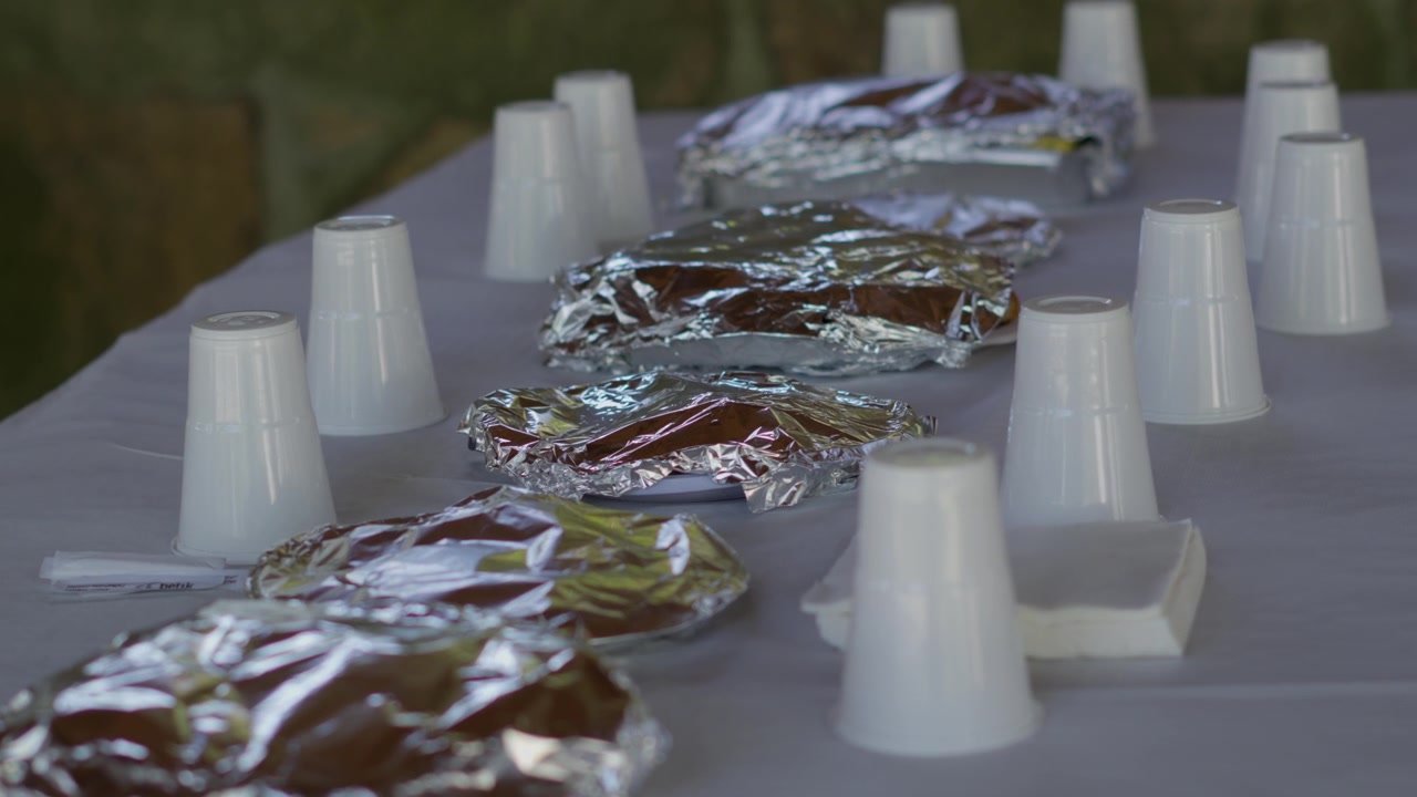 Vasos de plástico y platos envueltos en papel de aluminio cuidadosamente dispuestos sobre un mantel blanco al aire libre, creando un ambiente listo y acogedor para un picnic o evento.