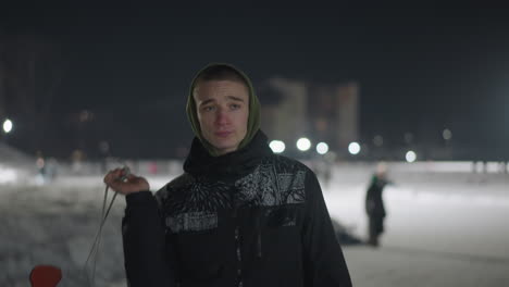young man in hooded jacket walking away during cold winter night with visible breath in air while holding skate boot over shoulder, soft lights and snowy path behind creating calm atmosphere