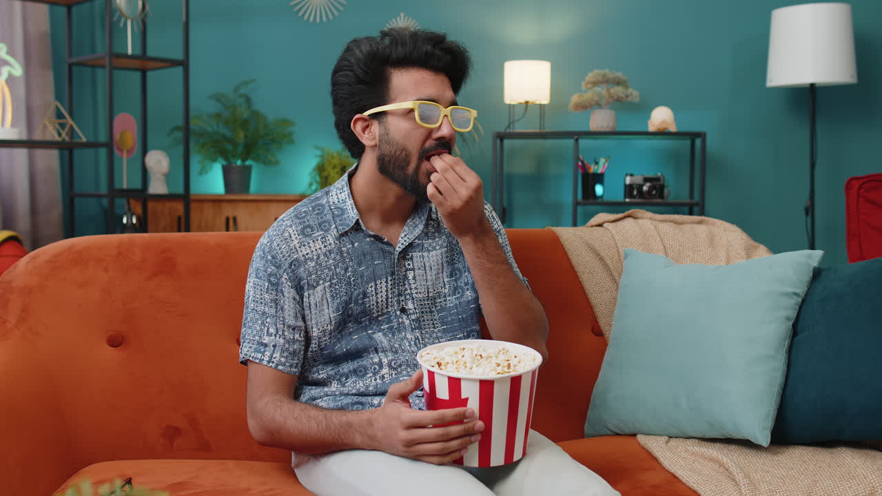 Indian man sits on sofa eating popcorn and watching interesting tv serial sport game online at home