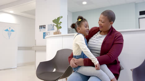 happy african american mother holding daughter on knees in hospital waiting room, slow motion
