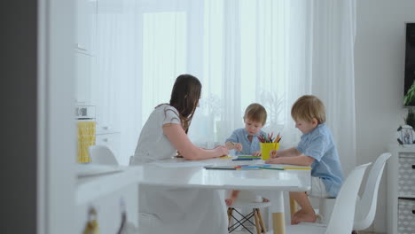 Mamá-Y-Sus-Dos-Hijos-Sentados-En-La-Mesa-De-La-Cocina-Dibujando-Lápices-De-Colores-Dibujando-En-Familia-En-El-Césped-En-El-Verano.-Los-Niños-Aprenden-A-Dibujar-Y-Mamá-Les-Ayuda-A-Dibujar.