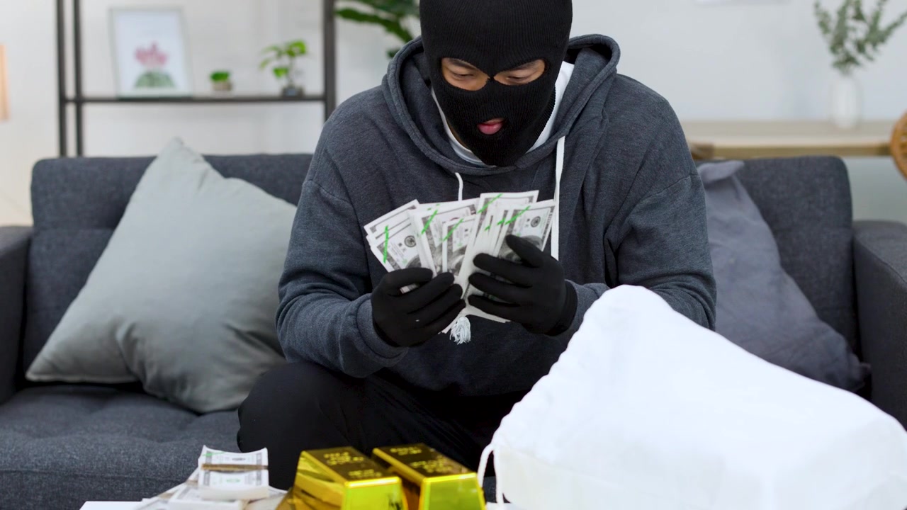 A masked criminal in a hoodie counts cash and gold bars after a successful burglary