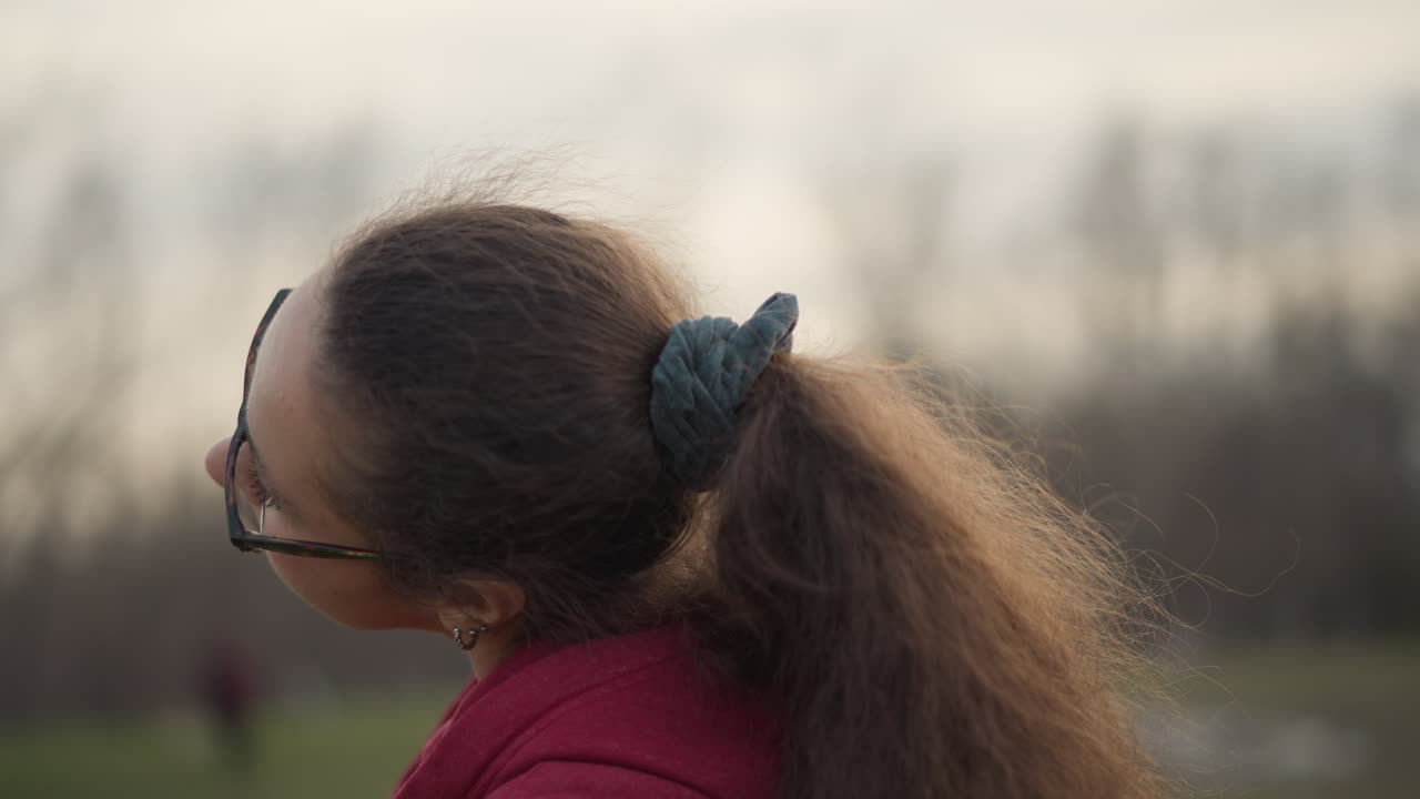 Young Woman Neck Stretch In Park, Red Hoodie And Glasses Performing Slow Neck Rolls And Gentle Head Tilts, Scrunchie Tied Ponytail, Overcast Suburban Field Background Conveying Calm SelfCare