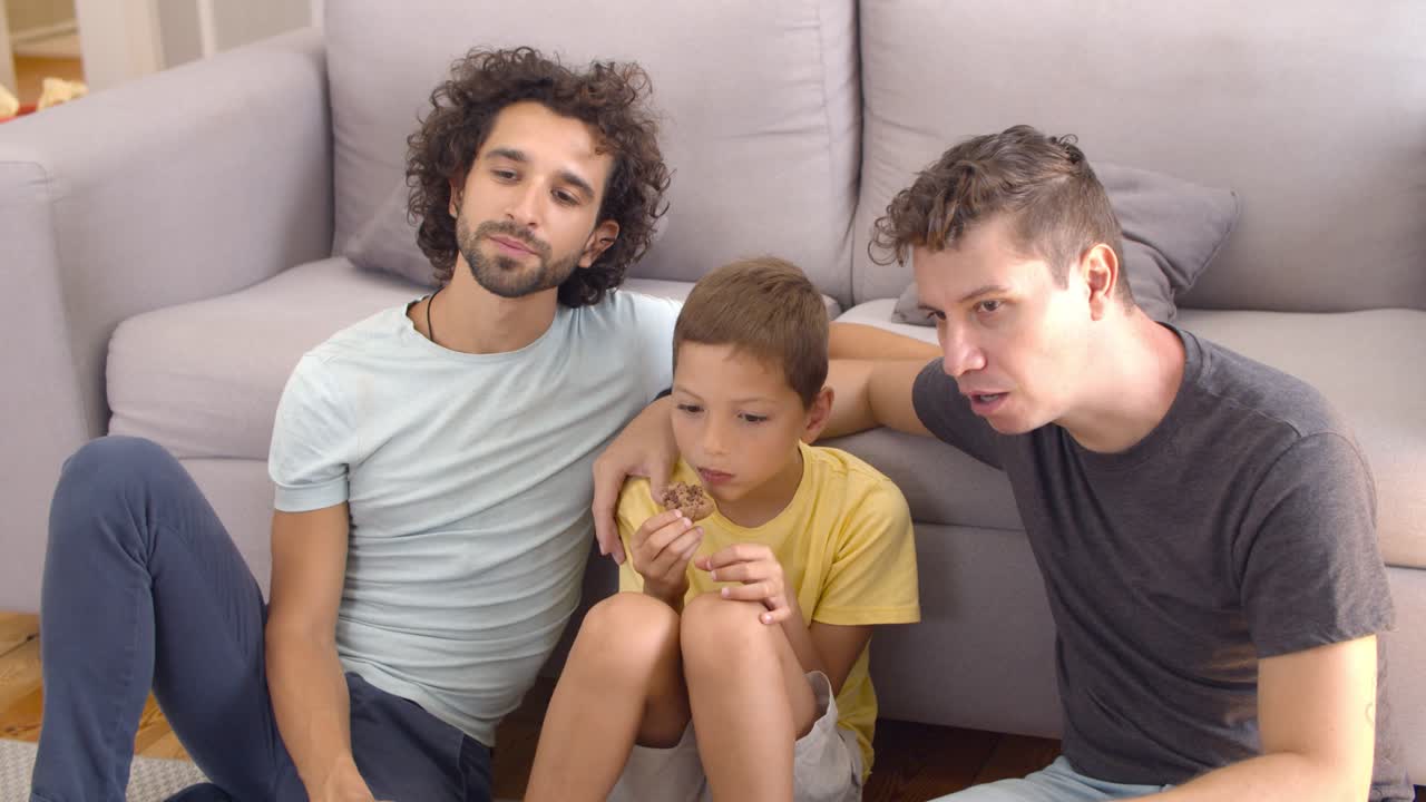 Genitori gay rilassati e bambino che mangiano biscotti e guardano un film.