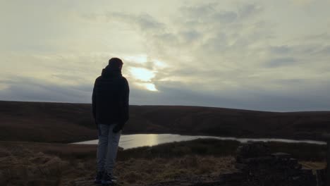 Young-boy-standing-and-looking-out-on-to-the-winter-landscape,-with-moorlands,-fens,-a-lake-and-stormy-winters-sky