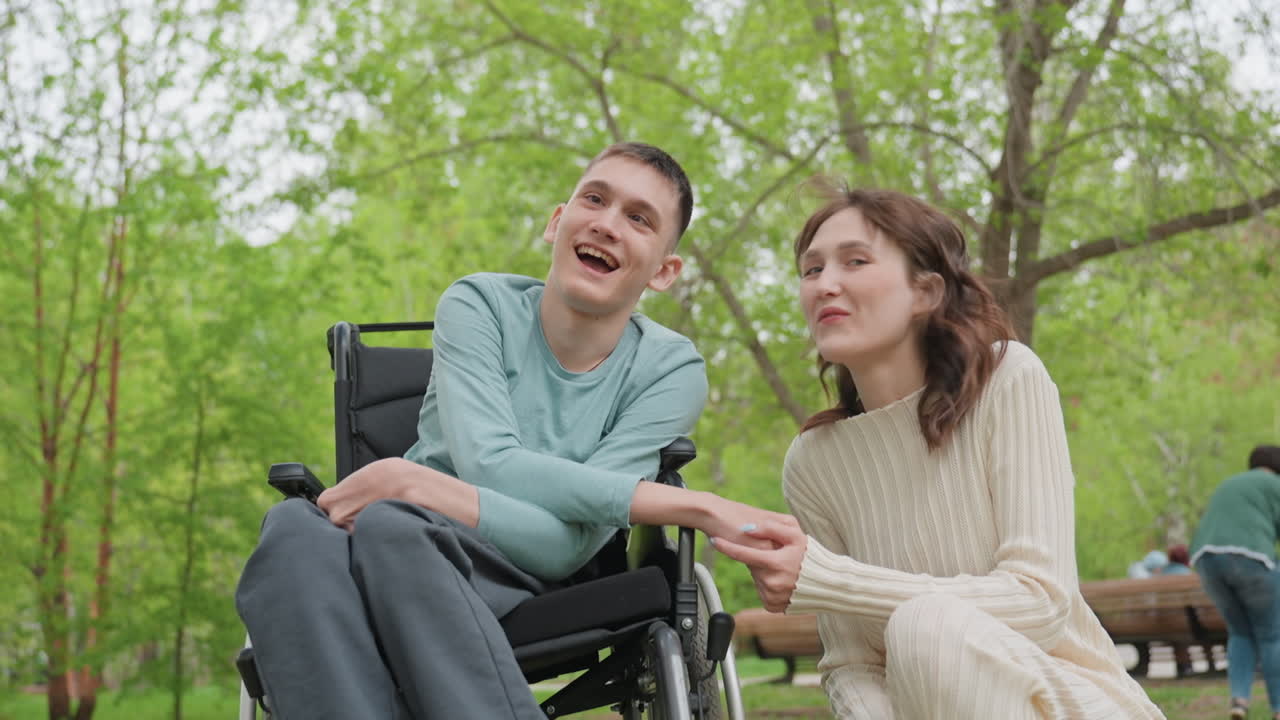 Young Man In Wheelchair Holding Hands With Companion In Park, Gentle Supportive Interaction On Bench Amid Green Trees, Sincere Smiles And Calm Conversation, Spring Sunlight, Inclusive Friendship