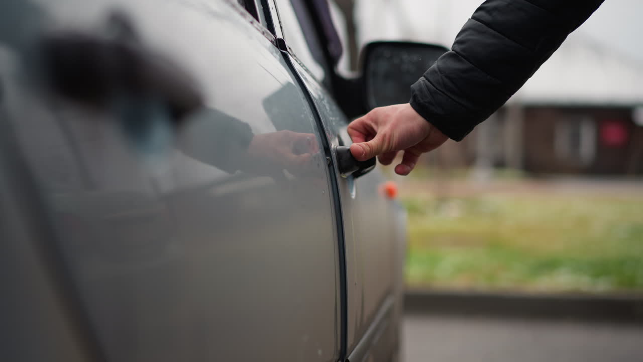 Side view of motorist opening door handle on ash coloured car with metallic finish and water droplets and snow on surface during winter day on urban street lined with parked vehicles