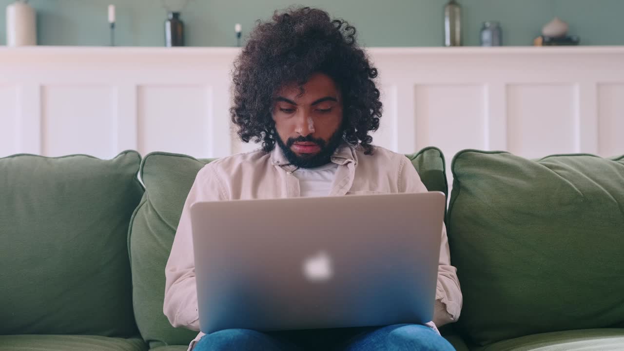 Young happy arabian man with laptop raises hands overjoyed sits on couch