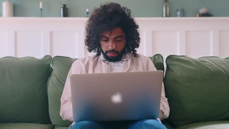 young happy arabian man with laptop raises hands overjoyed sits on couch
