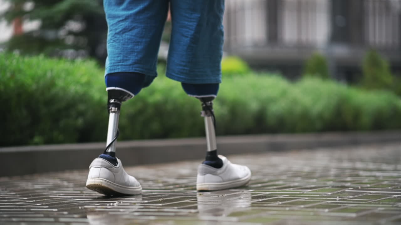 View of a walking man with prosthetic legs and white sneakers at rainy weather, greenery on the background