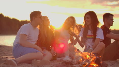 dos hombres y tres mujeres jóvenes están sentados alrededor de una fogata en la playa con cerveza. una de las mujeres está contando una historia interesante y divertida al atardecer en la noche de verano en la costa del lago.