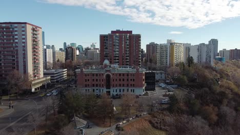 aerial-fly-over-cliffs-edge-forest-park-by-a-vintage-historic-commercial-building-next-to-densily-populated-high-rise-condominiums-apartment-buildings-hotels,-hospital-and-offices-in-Edmonton-Canada