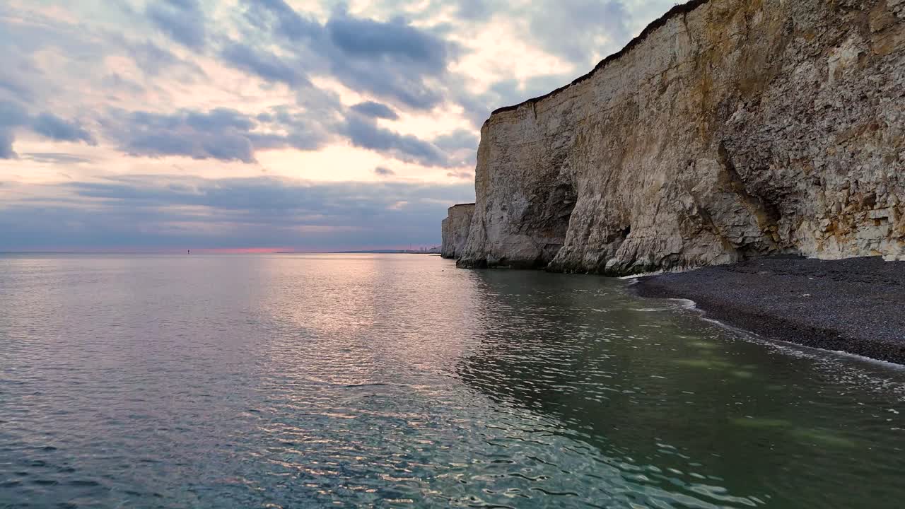 Sweeping drone view over Peacehaven’s pebbled beach and white cliffs in Sussex, with coastal caves below and Brighton visible in the distance. A stunning aerial shot of the English coastline.