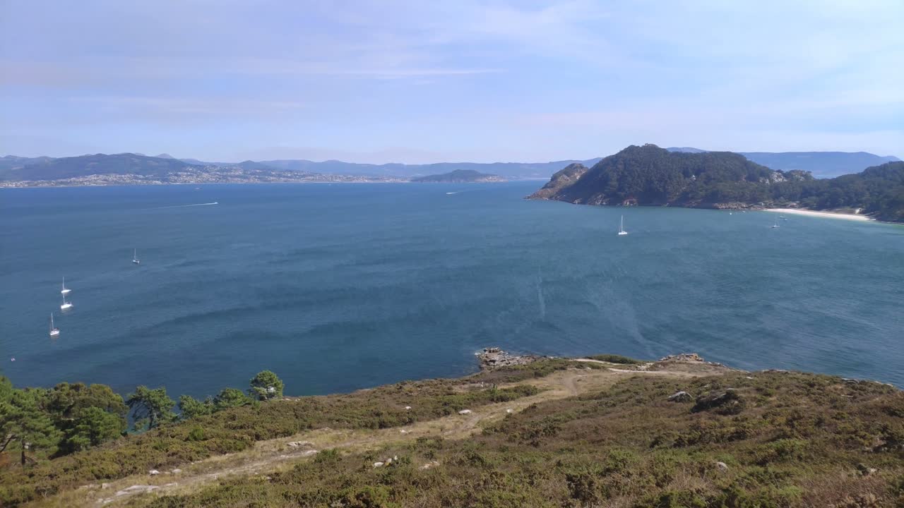 Elevated view of the sea and mountainous island with beach and boats sailing, sunny day, panoramic overhead shot turning right, Cíes Islands, Pontevedra, Galicia, Spain