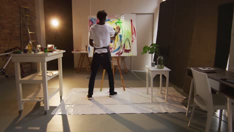 African-american-male-artist-wearing-apron-painting-with-paint-brush-on-canvas-at-art-studio