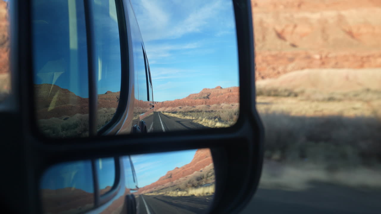 Ángulo desde el espejo retrovisor de una furgoneta camper con vista al desierto, la carretera y cielos azules durante un viaje por carretera a través del país
