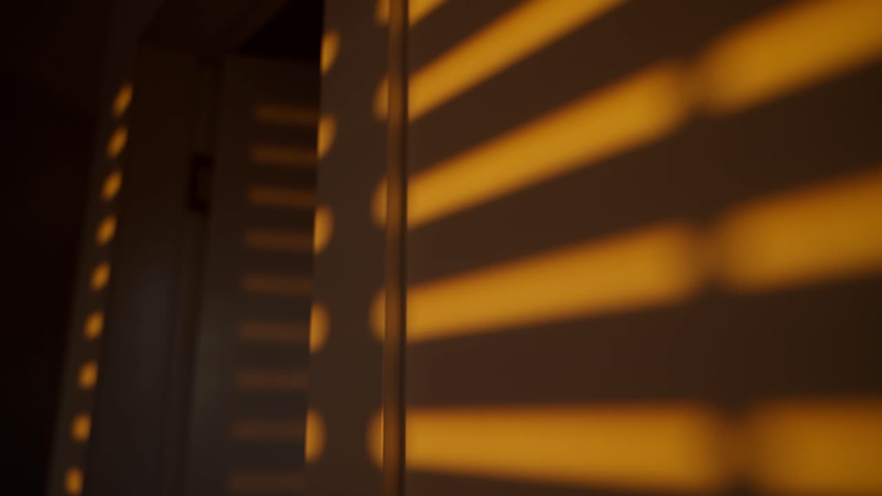 Shadow of mechanical rolling window blinds or shutters rolling down on wall. Orange colored sunlight in afternoon or early morning shining through window cracks as they shut down creating shadows