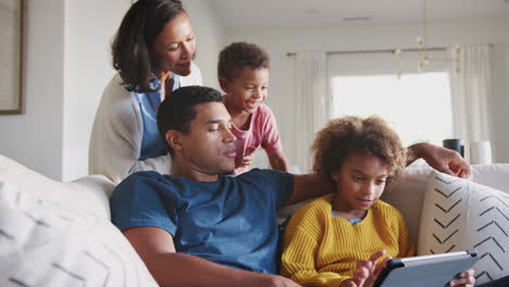 Dad-and-daughter-on-sofa-using-tablet-computer,-mum-standing-behind-them-holding-toddler,-low-angle