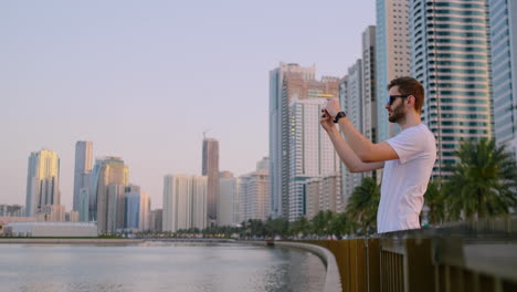 Glücklicher,-Gutaussehender-Mann-Mit-Sonnenbrille-Und-Weißem-T-Shirt-Und-Bart,-Der-Fotos-Mit-Einem-Smartphone-Macht,-Während-Er-Am-Wasser-In-Der-Sommerstadt-Im-Hintergrund-Und-In-Den-Gebäuden-Steht