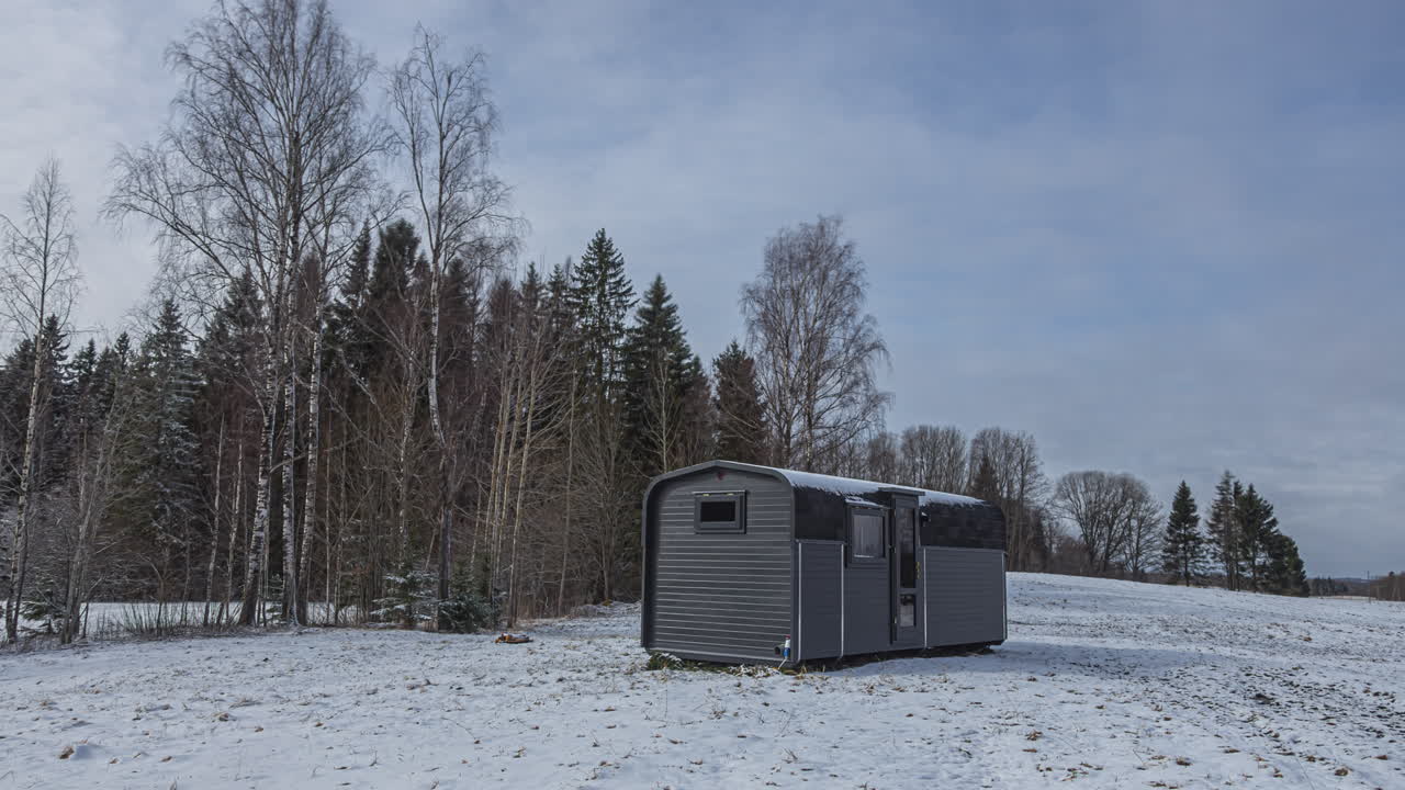 tiro de lapso de tiempo del frío día de invierno en un paisaje rural con nieve derretida del campo y nubes oscuras voladoras en el cielo - hora azul con amanecer gris y cabaña de madera