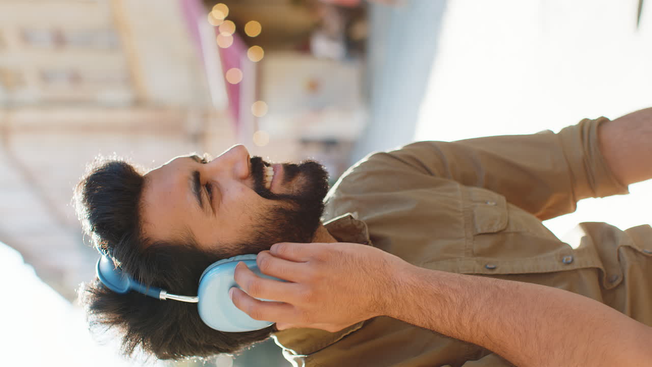 Happy young man in wireless headphones choosing listening music dancing outdoors city street