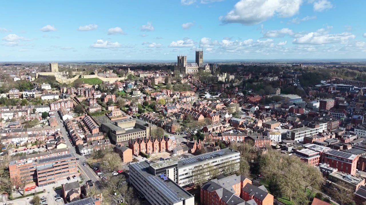 Ripresa aerea con drone della Cattedrale di Lincoln che si erge sopra un paesaggio urbano storico con edifici tradizionali, strade e un cielo nuvoloso luminoso in Inghilterra.