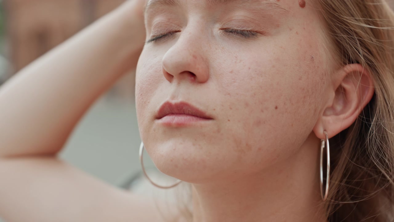 Giovane donna con lentiggini e orecchino a cerchio d'argento mostrata in primo piano mentre un'altra donna le acconcia i capelli. La scena si concentra sul profilo del suo viso, catturando movimenti sottili come un leggero aggiustamento