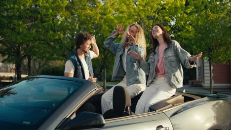 happy trio of young friends in denim jackets. a guy and two girls are dancing on the street near their gray convertible