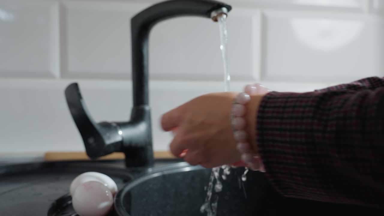 primer plano de las manos de la mujer abriendo el grifo de la cocina, recogiendo un huevo y lavándolo bajo agua corriente, huevos blancos frescos descansan en el fregadero negro mientras fluye el agua