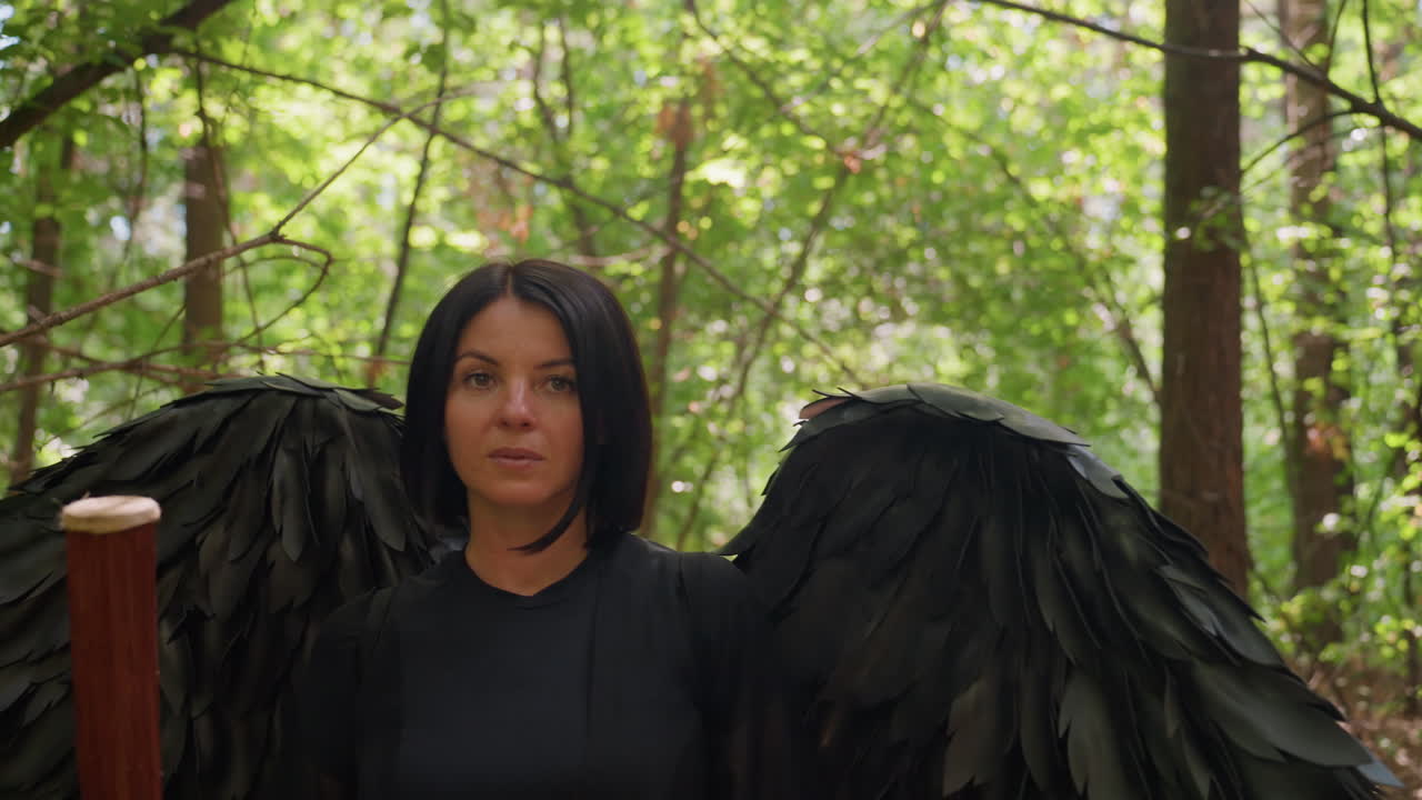 Rear view of actress dressed as black fairy walking gracefully through forbidden woodland with staff, sunlight filtering through trees as she turns with serious face, surrounded by magical forest