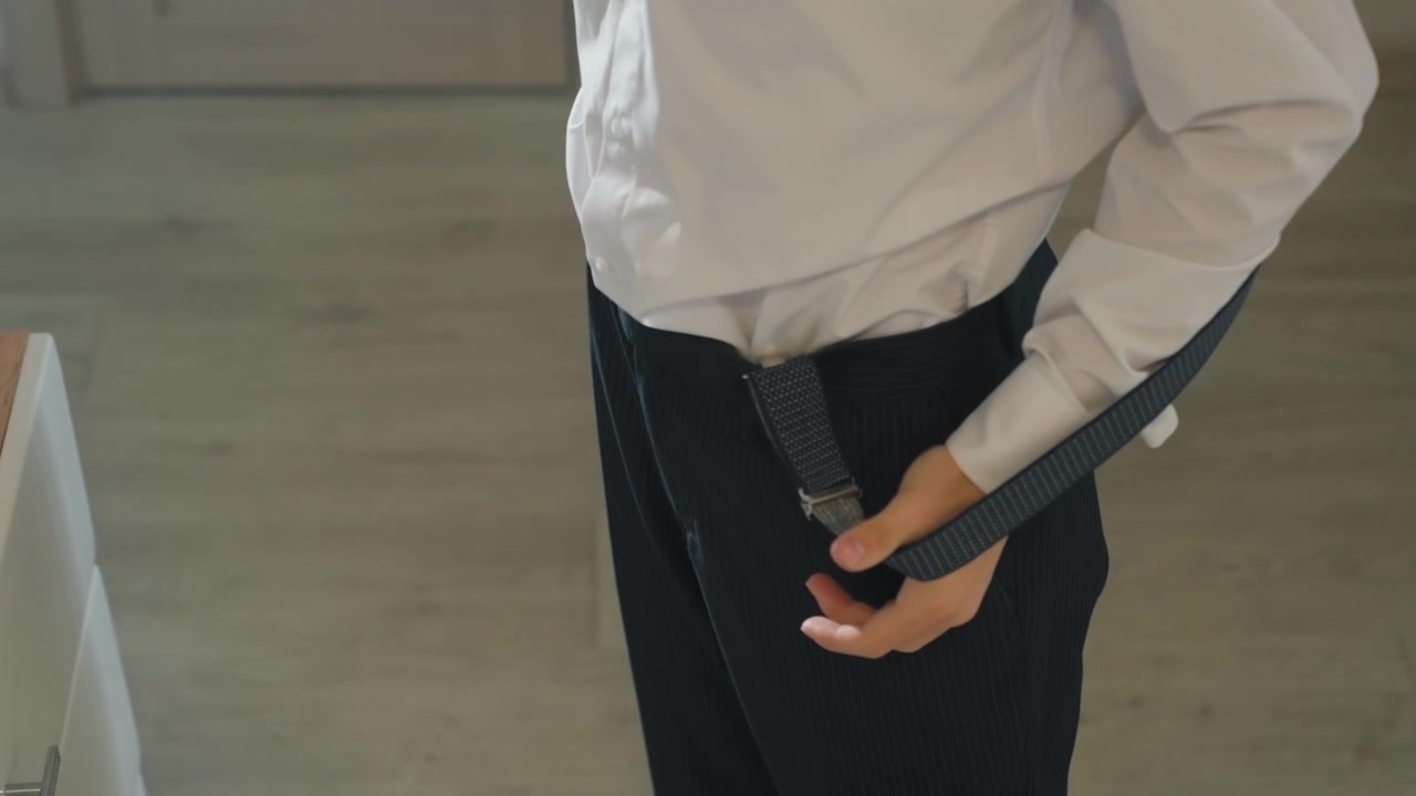 Asian man adjusting suspenders and shirt, preparing tailored trousers in sunlit apartment, focused hands smoothing navy textured fabric, neat white collar, calm morning ritual before professional