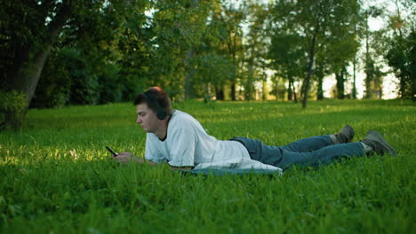 young man lying on grass with headphones on, engrossed in his phone, surrounded by lush greenery and tranquil park environment, his relaxed posture and focused gaze reflect leisure