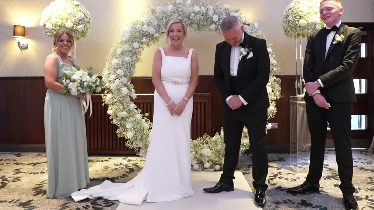 Cerimonia di matrimonio al coperto in Scozia con una sposa e uno sposo felici in piedi sotto un arco floreale, con damigella d'onore e testimone dello sposo presenti, che cattura un momento di amore, celebrazione e impegno.