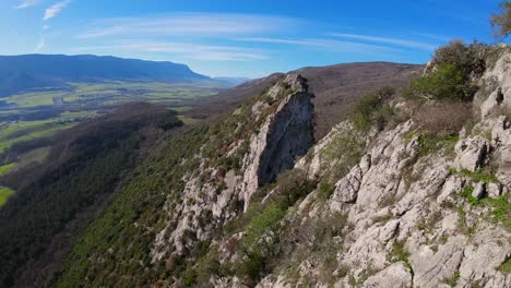 FPV-drone-aerial-image-taken-from-the-top-of-a-towering-stone-mountain,-a-calming-natural-image-with-shades-of-green,-grey,-and-blue-sky