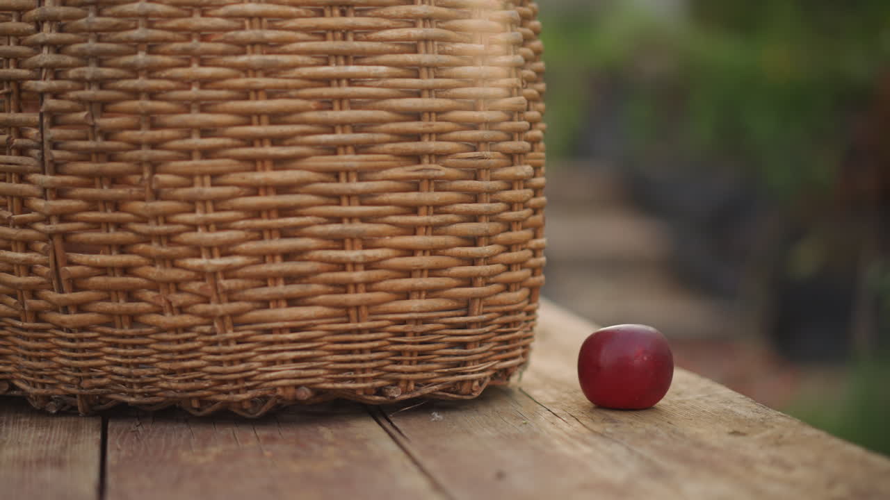 Cesta de vime com maçã vermelha sobre mesa de madeira, fundo bokeh suave, cena rústica de jardim ao ar livre, luz suave da manhã, foco na pequena fruta ao lado da cesta trançada, atmosfera de colheita tranquila.