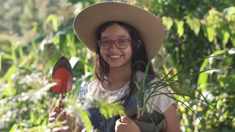 niña sonriente con sombrero y gafas, de pie en el medio de un jardín botánico con una pala y una planta de cebolla en sus manos