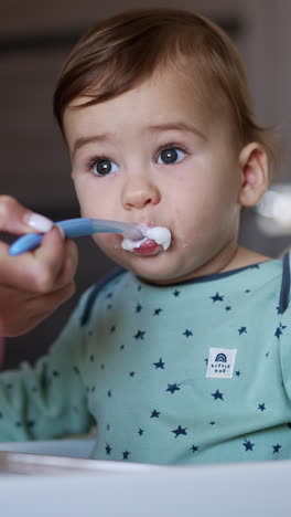cute caucasian kid in blue shirt sitting at the feeding table. mom gives porridge to her son and cleans his mouth with the spoon. blurred backdrop. vertical video