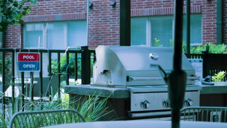 las ondas de calor surgen de una parrilla de barbacoa de acero inoxidable cerca de un letrero de piscina abierta y una puerta de hierro forjado, respaldada por un edificio de ladrillo y con una mesa de picnic en primer plano