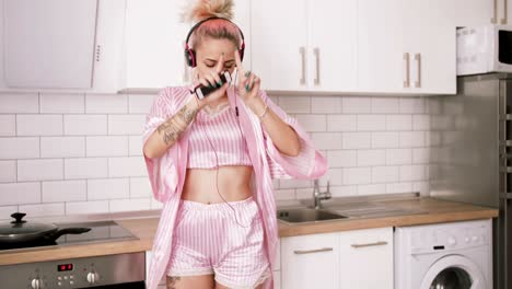 happy young woman with pink hair dancing in kitchen wearing pink pajamas and listening to music with headphones