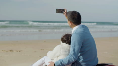 back view of a man holding smartphone and taking selfie while hugging his lovely daughter at the beach