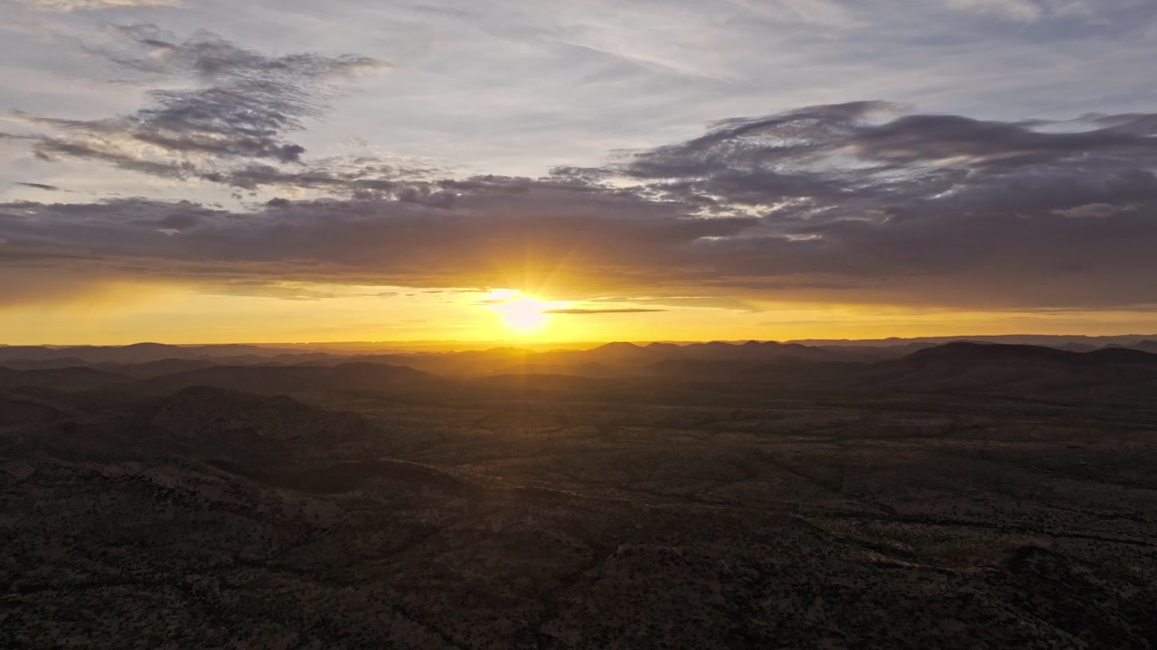 Vista aérea do nascer do sol sobre o deserto da Namíbia, um brilho dourado quente se espalhando ao longo do horizonte sob um céu nublado suave e em camadas.
