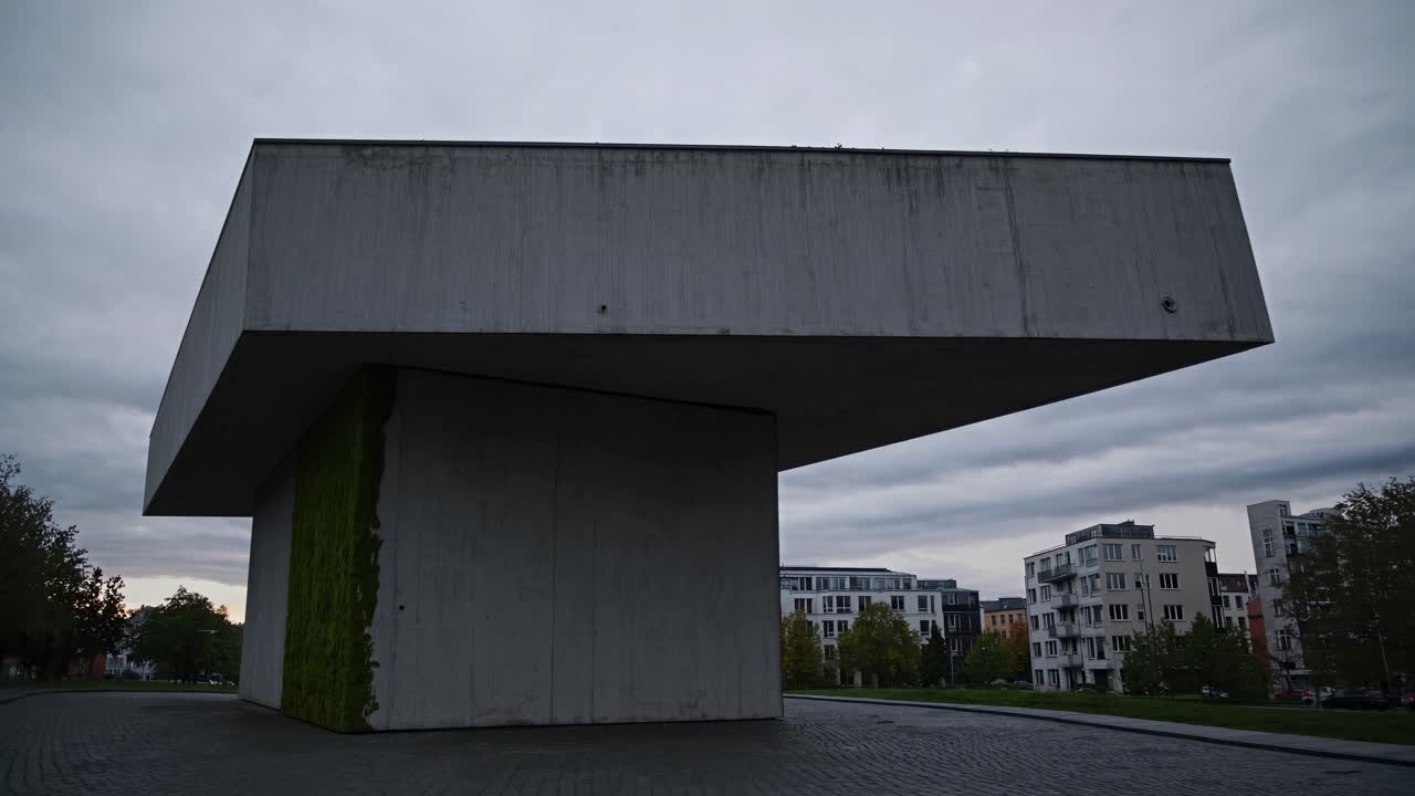 Concrete structure with moss growing on one side stands tall against a backdrop of residential buildings and a cloudy sky, showcasing the unique blend of nature and urban design in Berlin