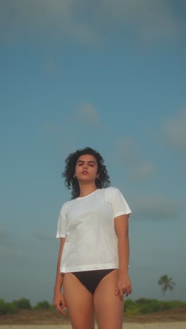 indian woman in white t-shirt and bikini bottoms stands against blue sky in low-angle beach shot