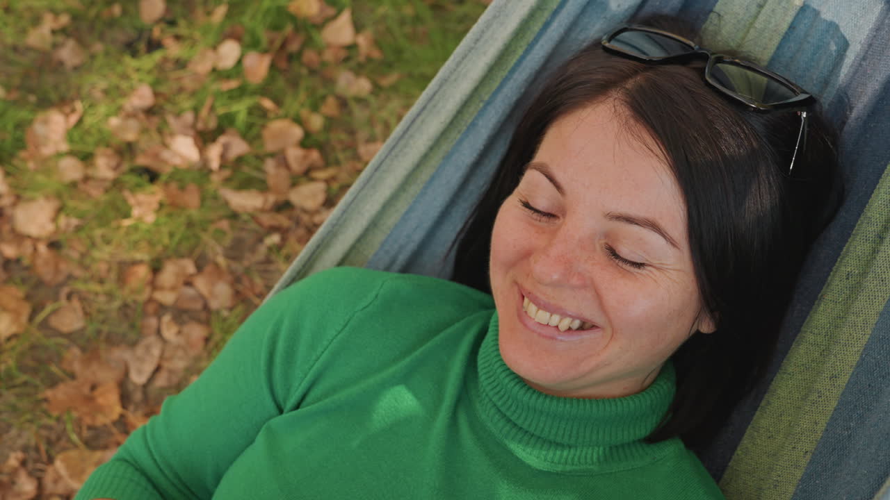 Donna caucasica in un'amaca da campeggio che legge un libro, uno sguardo giocoso da dietro la copertina e un sorriso caloroso, con un maglione a collo alto verde e occhiali da sole sulla testa, foglie autunnali sotto i piedi, un pomeriggio rilassato, una brezza leggera, in uno stato di consapevolezza.