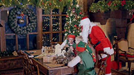 elves, children and santa claus in decorated room on holidays. little kids sitting at the table with elves and opening envelope. santa claus is standing near the children in his residence.