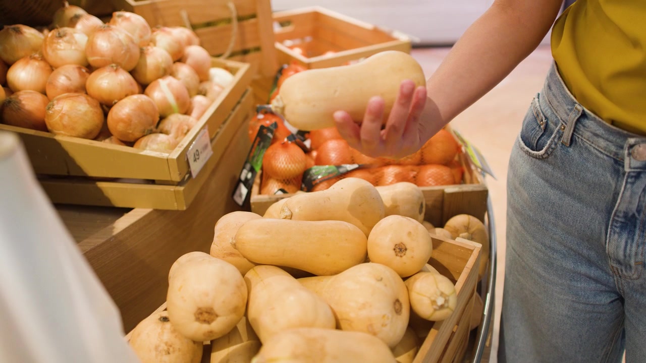A shopper holds and examines organic butternut squash at a supermarket vegetable display stand
