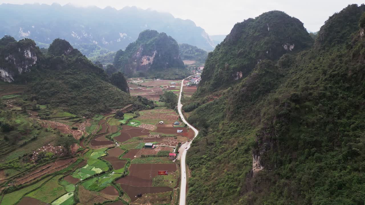 Pemandangan Vietnam Utara dengan pegunungan karst dan lahan pertanian pedesaan yang dibagi oleh jalan berkelok melalui lembah dengan desa kecil dan tanaman hijau subur, diambil dari pandangan drone udara.