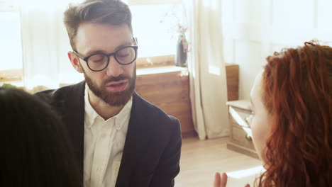 hombre serio con anteojos hablando con dos mujeres mientras se sienta en una habitación luminosa por la mañana