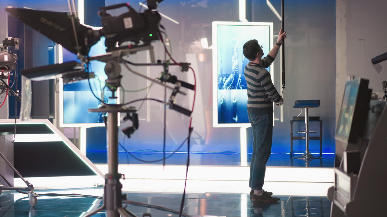 Man holding boom microphone, adjusting it during video production in modern studio, neon lighting, and crew equipment visible, professional sound recording process in cinematic environment